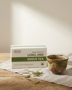 Box of Wu Wei Living Long Jing Green Tea with a ceramic cup on a wooden surface.