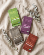 Four packages of Wu Wei Living loose leaf tea on a beige fabric background with eucalyptus leaves.
