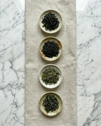 Four small ceramic bowls with different types of tea leaves on a textured mat against a marble background. — Wu Wei Living