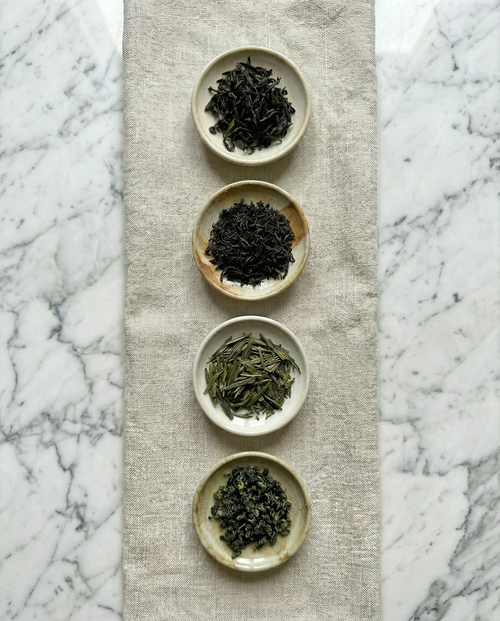 Four small ceramic bowls with different types of tea leaves on a textured mat against a marble background. — Wu Wei Living