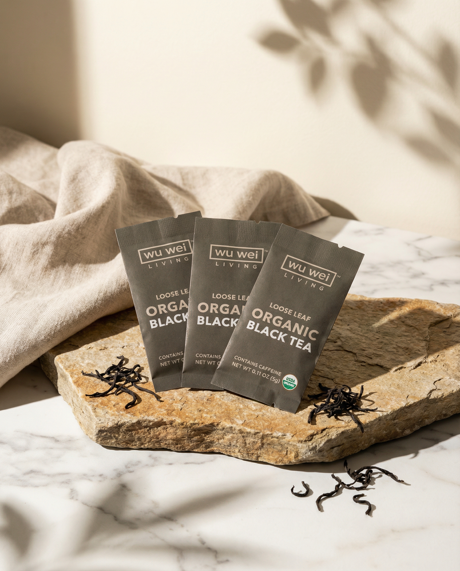 Three packages of Wu Wei Living Organic Loose Leaf Black Tea on a stone surface with tea leaves scattered around.