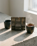 Wu Wei Tea organic black tea packages with steaming cups on a concrete surface.