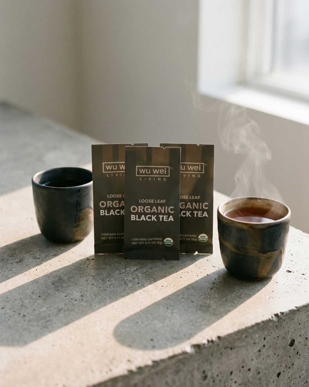 Wu Wei Tea organic black tea packages with steaming cups on a concrete surface.