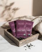 Three purple packages of Wu Wei Living Organic Loose Leaf Tea on a wooden tray with a neutral background.