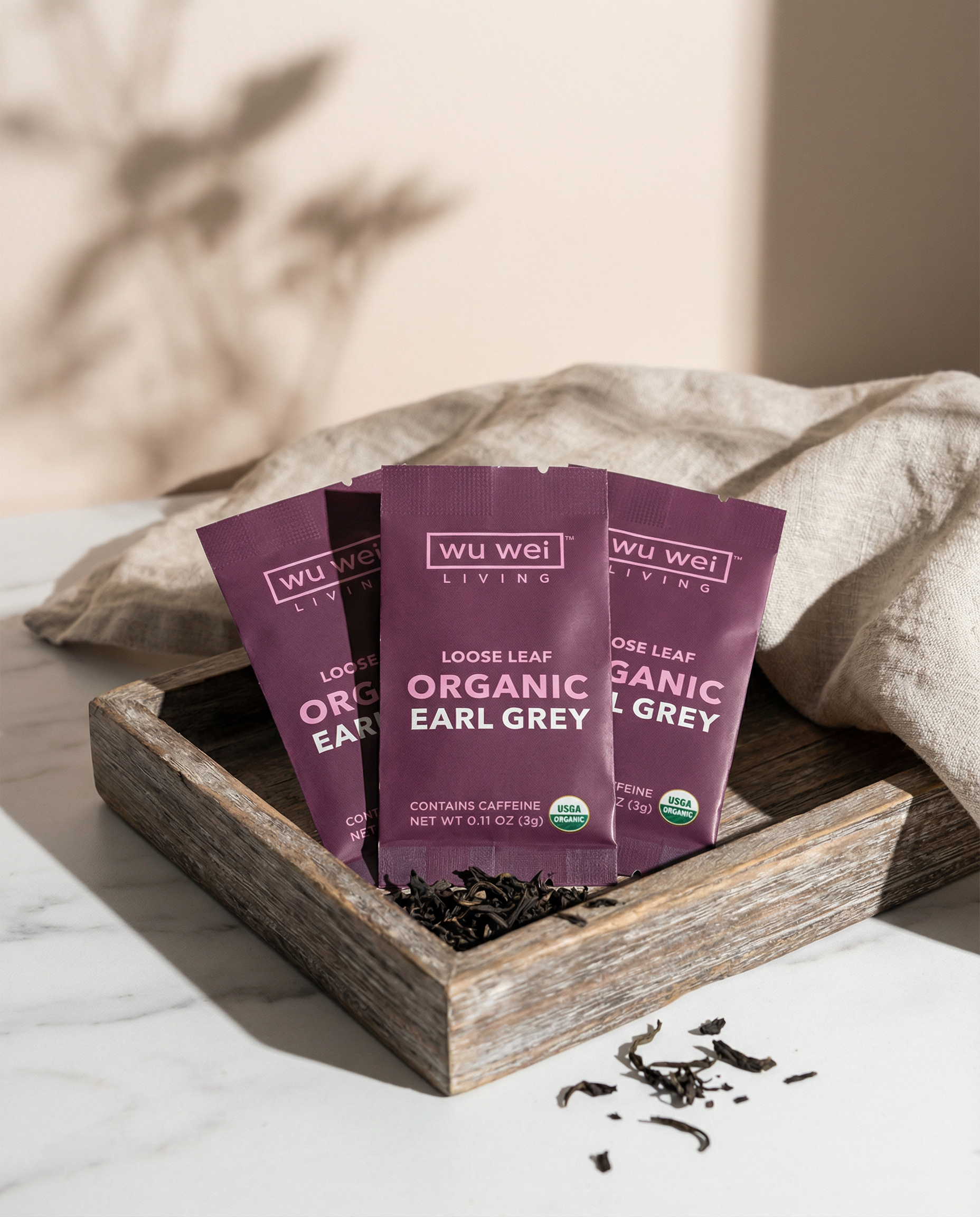 Three purple packages of Wu Wei Living Organic Loose Leaf Tea on a wooden tray with a neutral background.