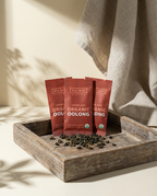 Three red tea packages on a wooden tray with loose tea leaves.- Wu_Wei_Living_Organic_Oolong